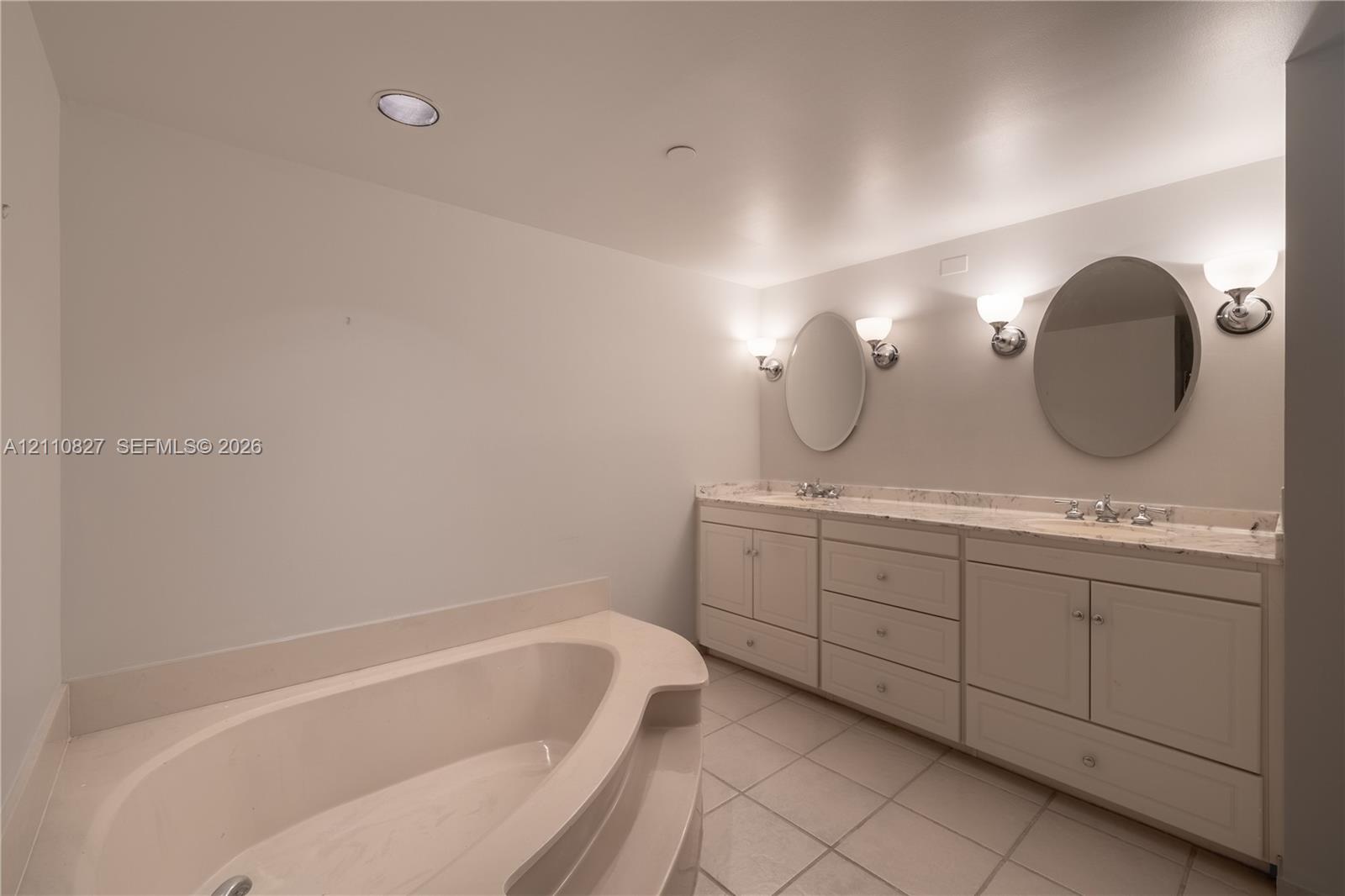 Dual-sink vanity with stone-surfaced countertop, drop-in soaking tub, tile flooring, and wall-mounted round mirrors