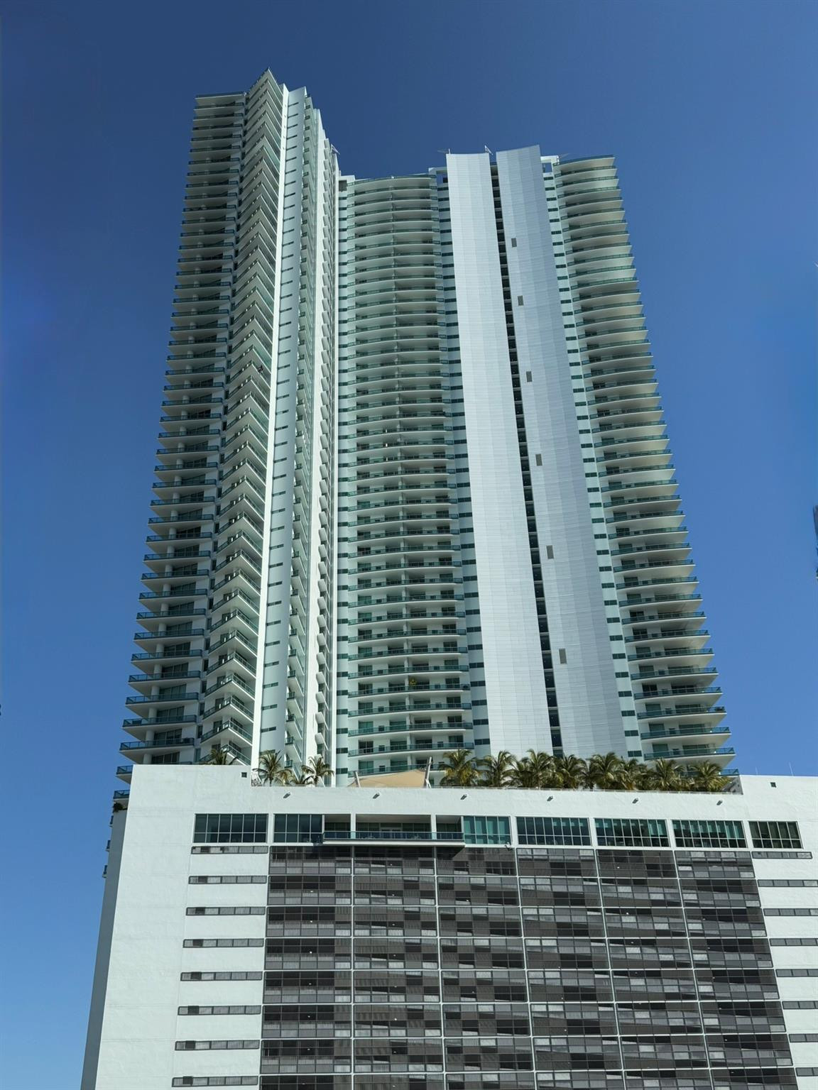 Contemporary high-rise building featuring a multi-story base, extensive window-lined facade, numerous private balconies, and a rooftop amenity deck wi