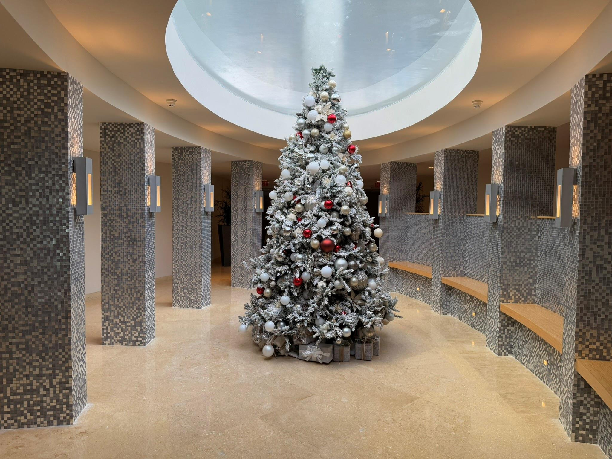 Open circular atrium featuring a skylight, light-toned polished flooring, mosaic-tiled columns, integrated wood benches, and wall-mounted sconces
