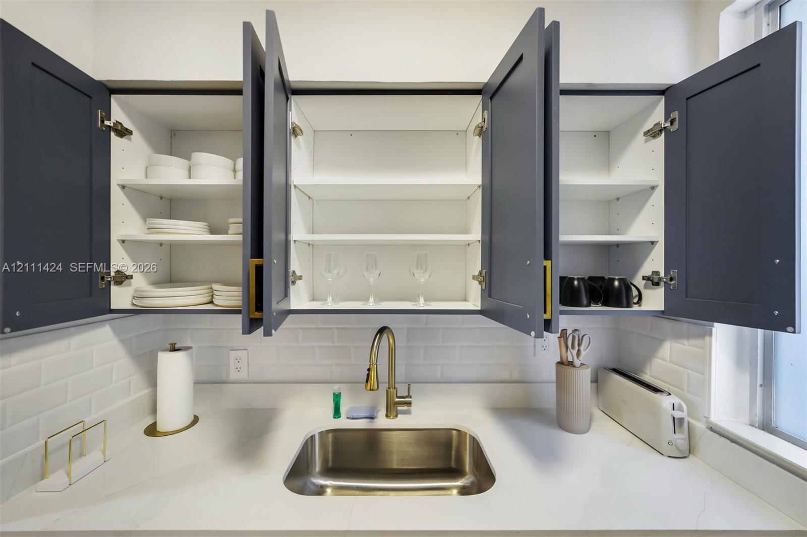 Shaker-style cabinetry with matte finish, white subway tile backsplash, solid-surface countertop, stainless steel undermount sink, and brass-finish go
