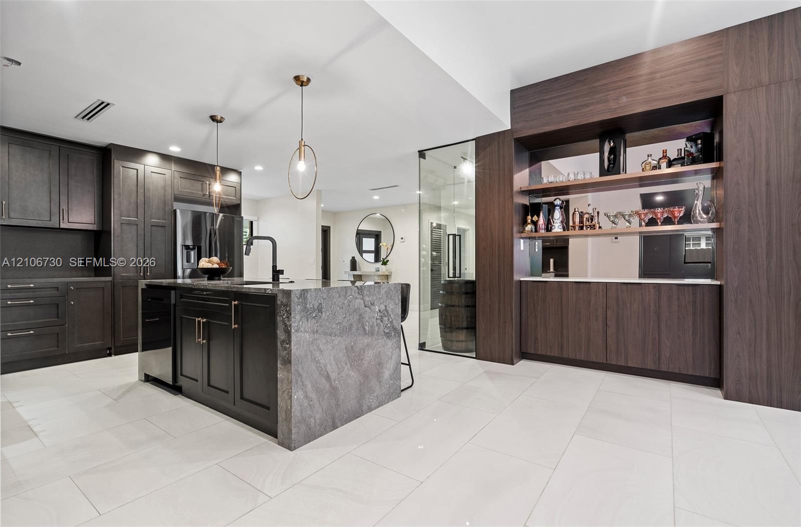 Kitchen featuring dark wood-finish cabinetry, a waterfall-edge stone island, built-in shelving with mirrored backsplash, and large-format tile floorin