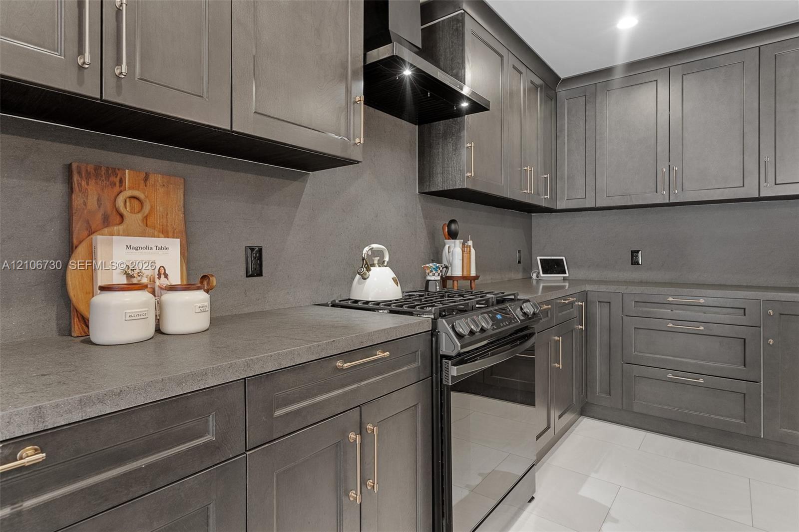 Kitchen featuring charcoal-toned cabinetry, solid-surface countertops with integrated backsplash, stainless steel gas range, black range hood, and lar