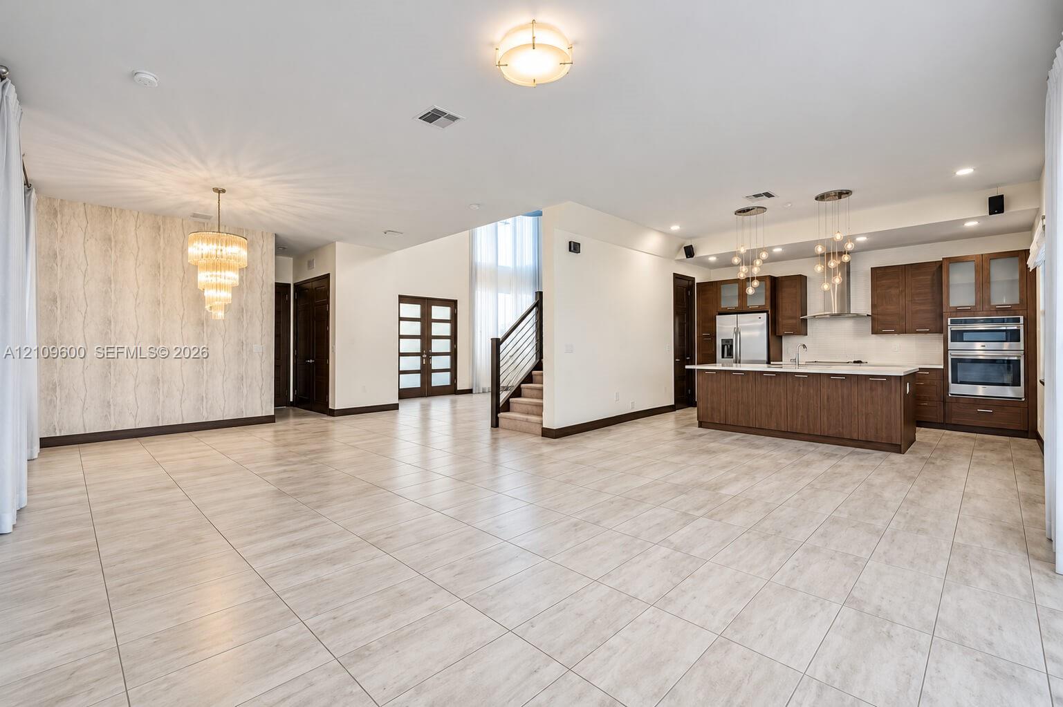 Open-concept interior featuring large-format tile flooring, wood-finish cabinetry, built-in stainless steel appliances, tiered crystal-style chandelie