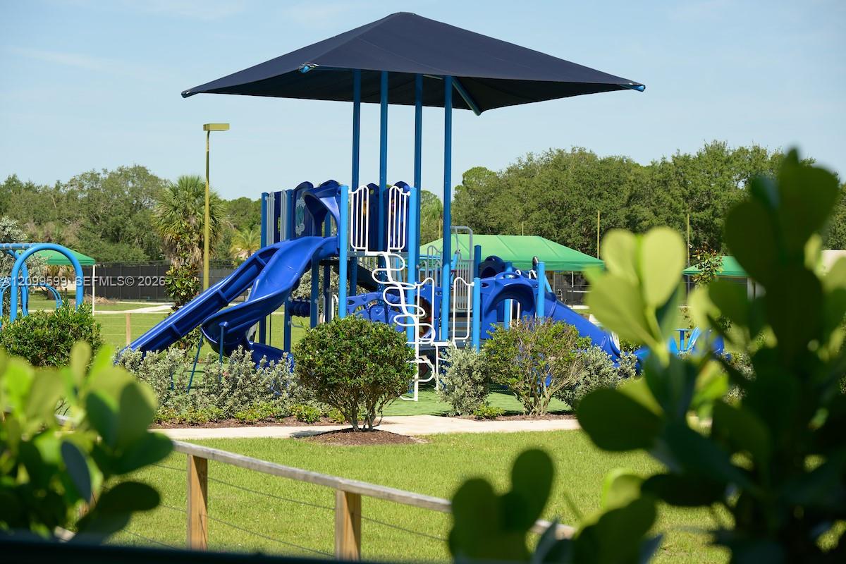 Outdoor recreational area featuring a playground structure with shade canopy, slides, and turf surfacing, enclosed by wooden post-and-rail fencing