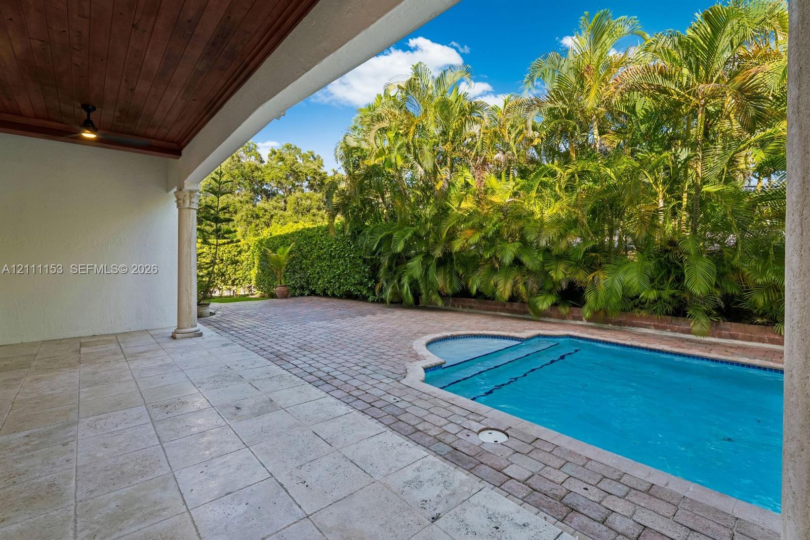 Covered patio with natural stone flooring and wood-paneled ceiling, featuring a swimming pool, brick paver decking, and perimeter landscaping