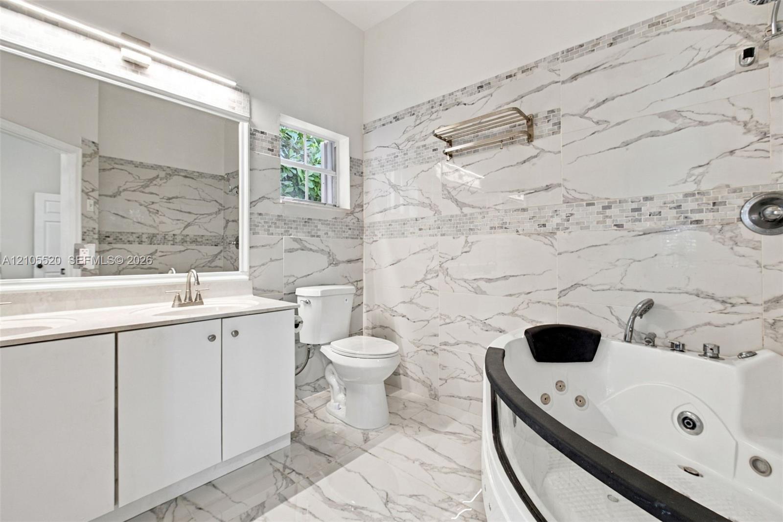 Bathroom featuring floor-to-ceiling large-format tile with stone-pattern finish, jetted soaking tub, white vanity cabinet with countertop basin, and o