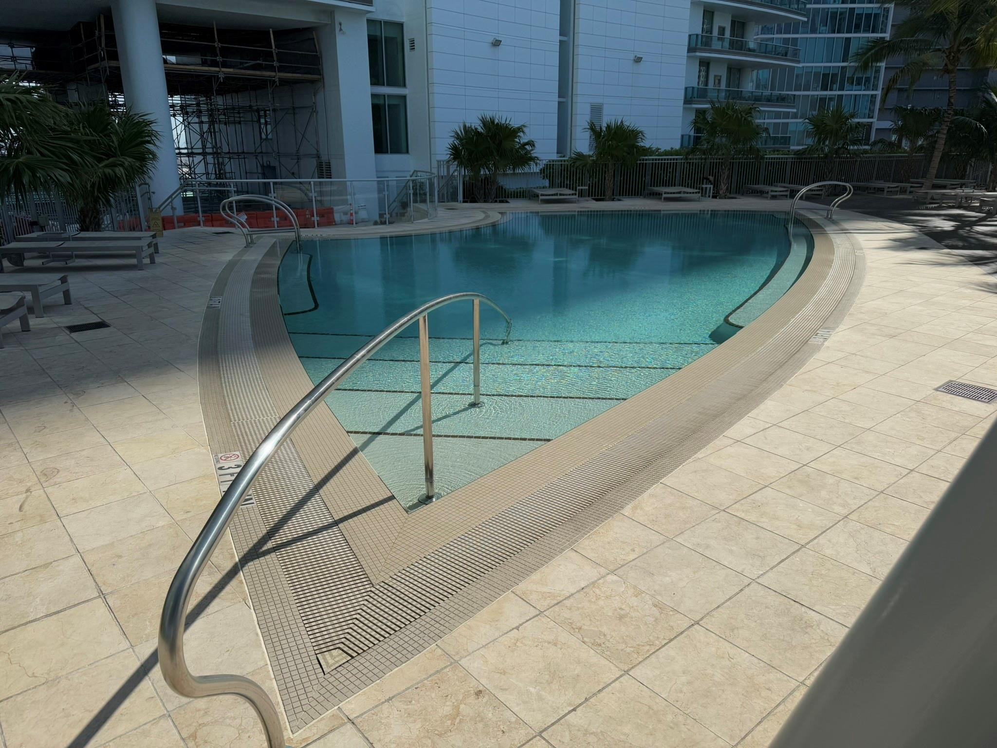 Curved swimming pool with stainless steel handrails, surrounded by light-toned tile decking